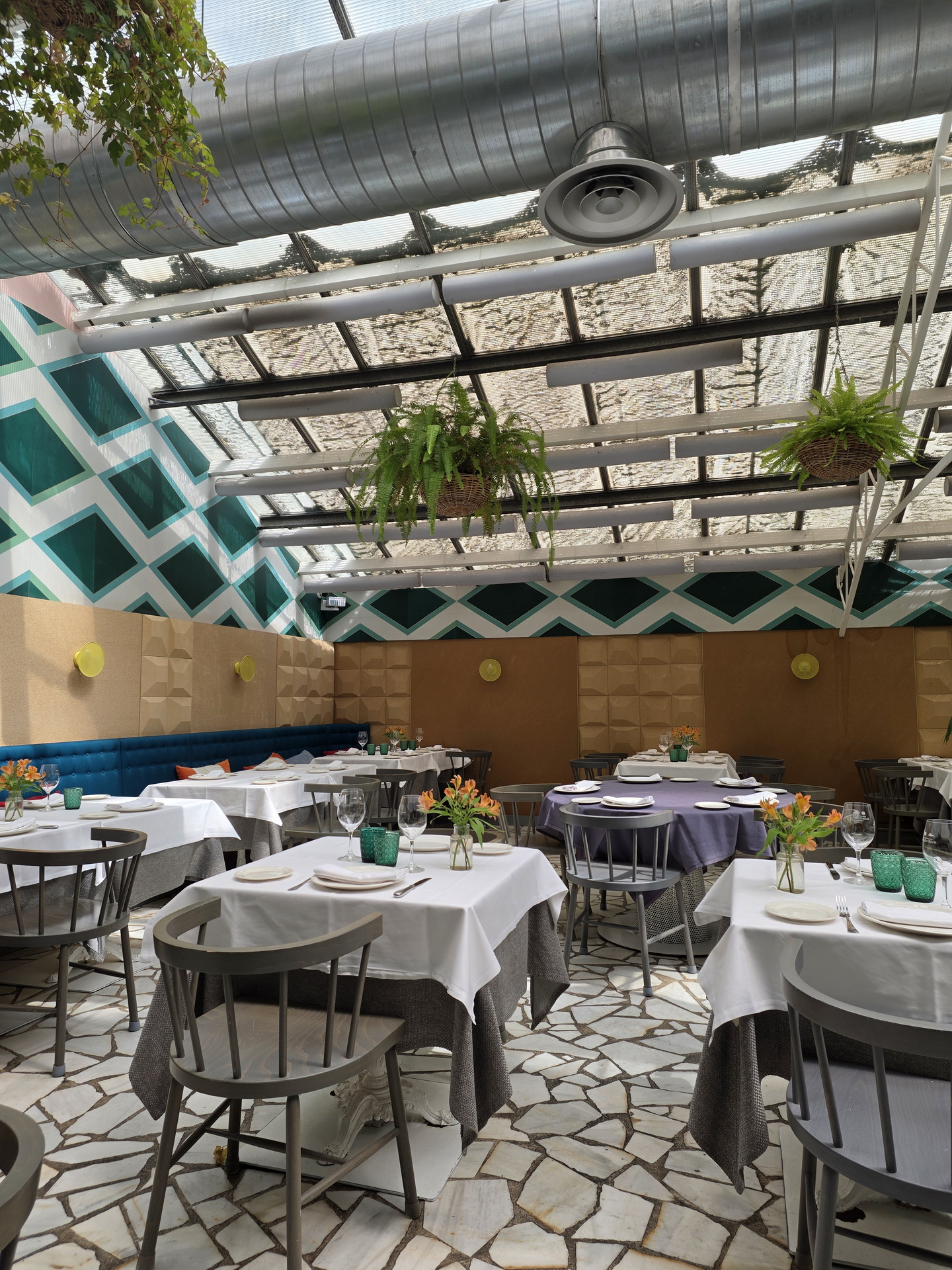 Restaurante con techo acristalado y mesas decoradas, sillas de madera, plantas colgantes y ambiente luminoso.