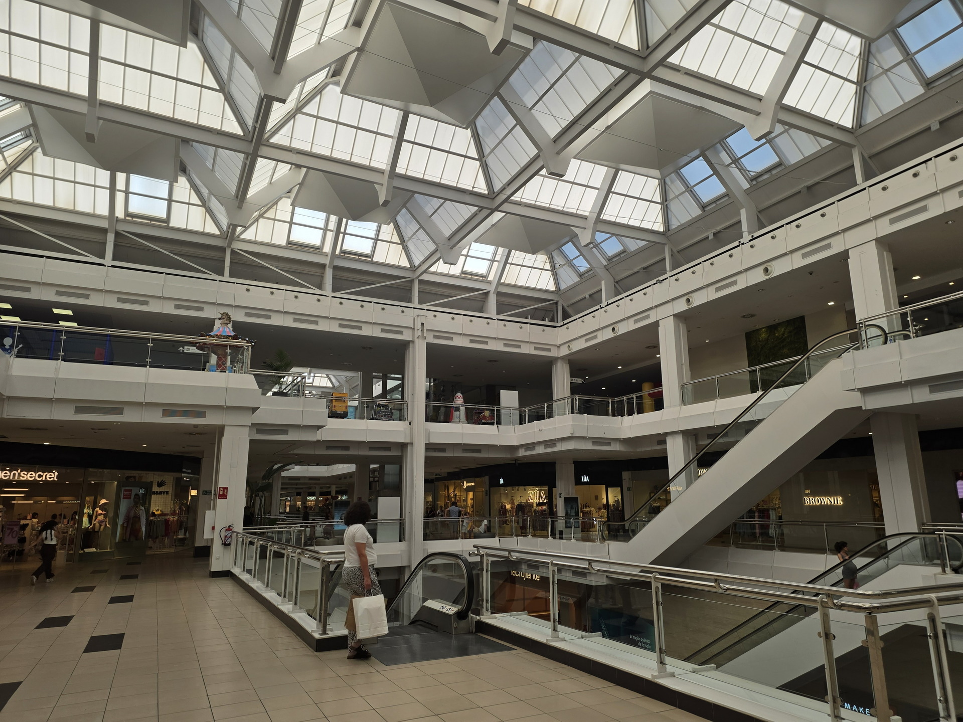 Centro comercial moderno con techo de vidrio y escaleras mecánicas, tiendas y personas caminando.