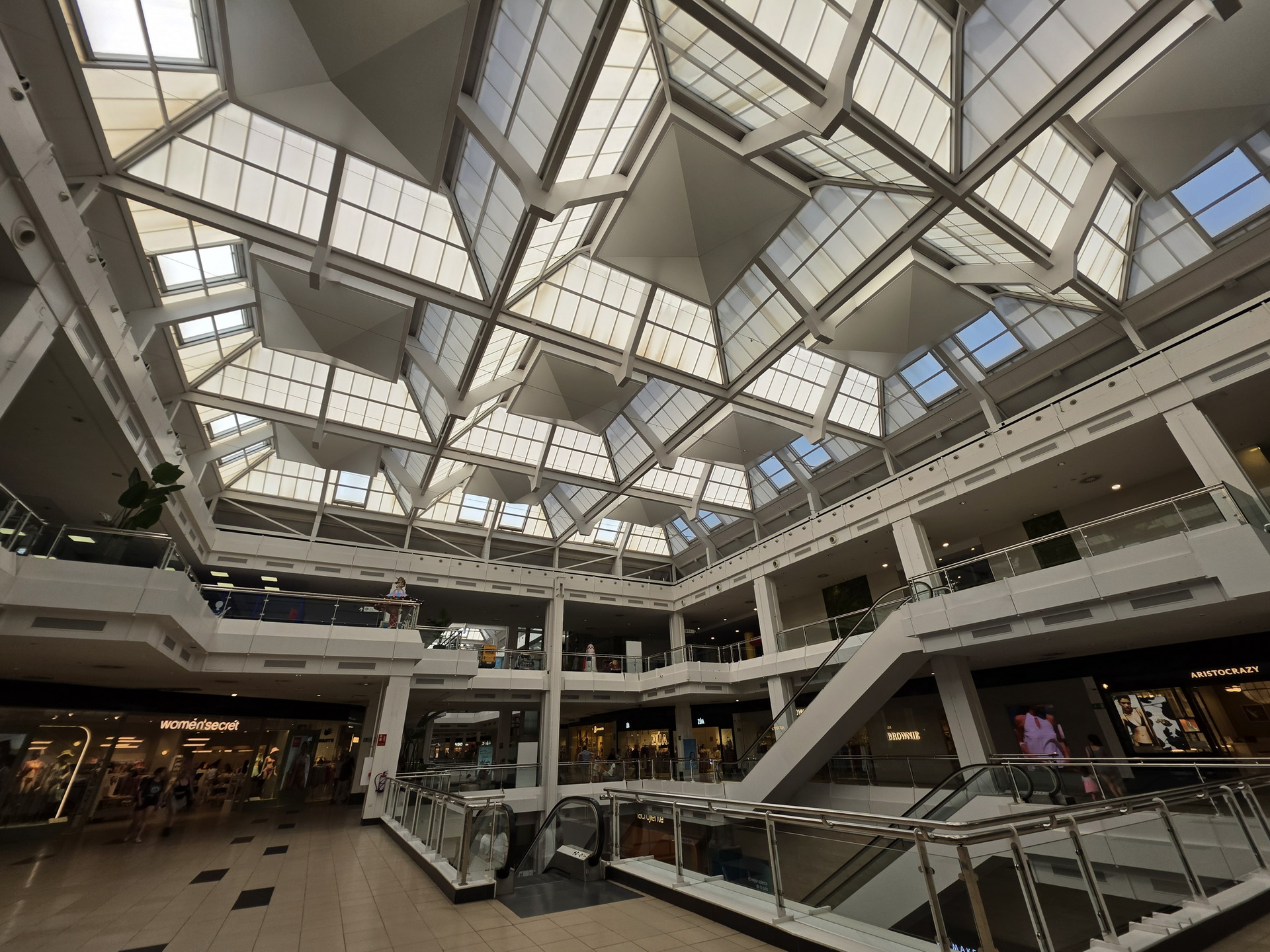 Centro comercial amplio con techo de cristal geométrico y tiendas como Women's Secret y Aristocrazy.