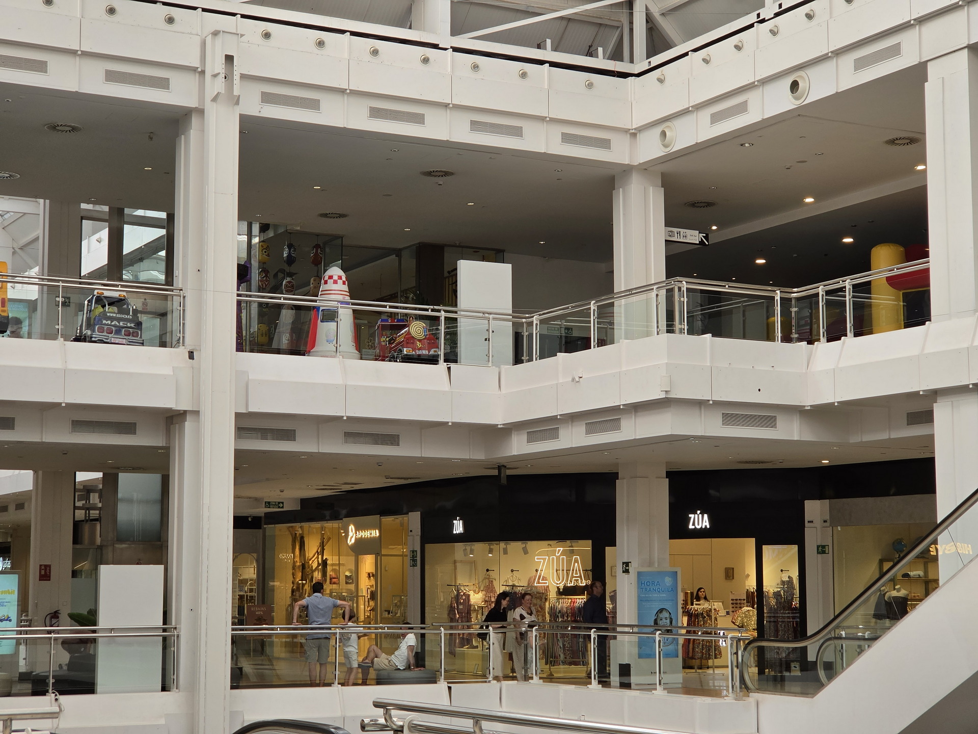 Centro comercial moderno con tiendas y personas caminando, escaleras mecánicas y decoración colorida en el segundo nivel.