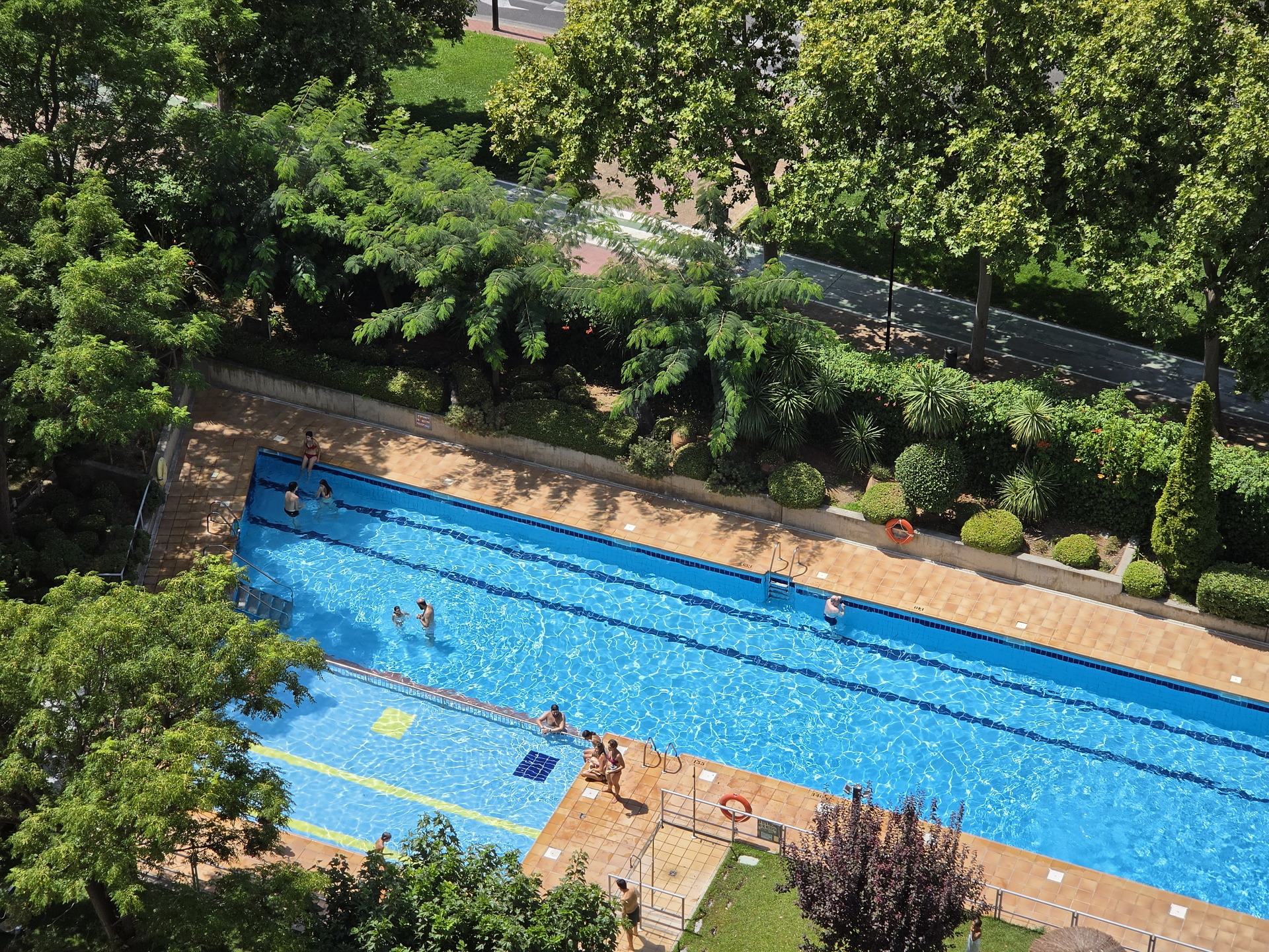 Vista aérea de una piscina al aire libre rodeada de árboles y personas disfrutando del agua en un día soleado.