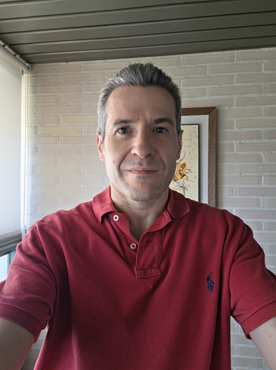 Hombre de mediana edad sonriendo, vistiendo polo rojo, junto a pared de ladrillos, luz natural entrando por la ventana.