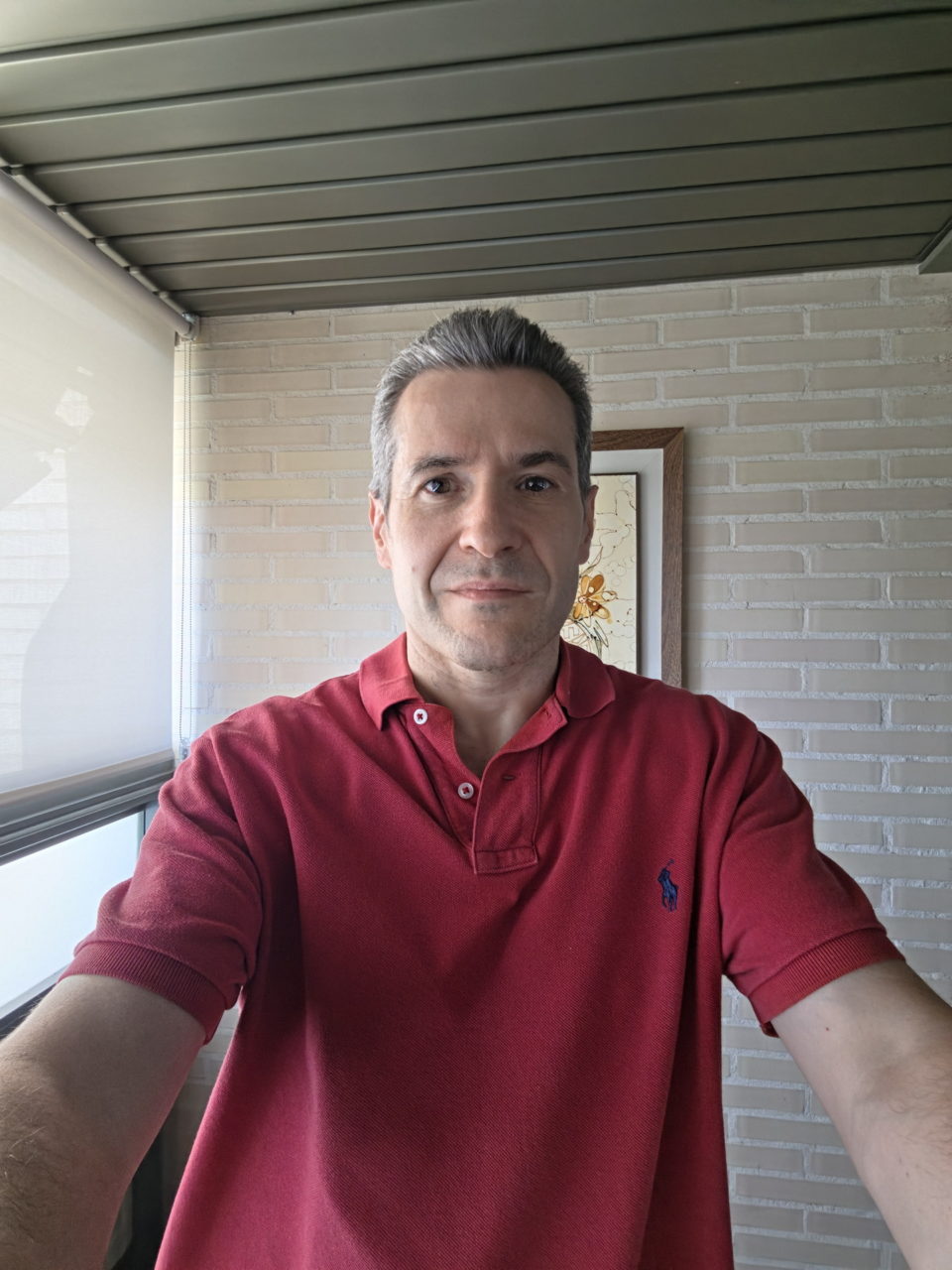 Hombre con polo rojo haciendo selfie en balcón con paredes de ladrillo blanco. Luz natural y decoración en el fondo.
