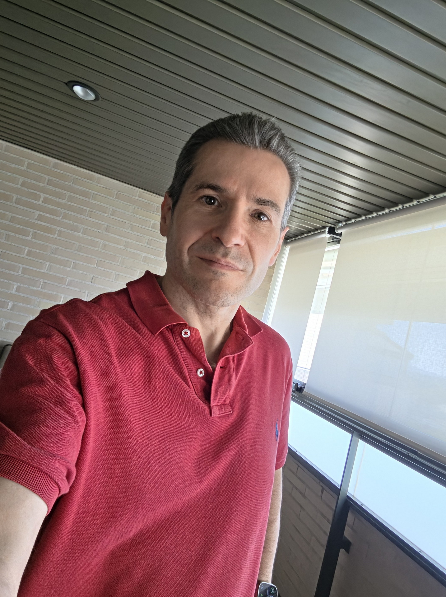 Hombre con camiseta roja en terraza iluminada con paredes de ladrillo y techo de listones metálicos.