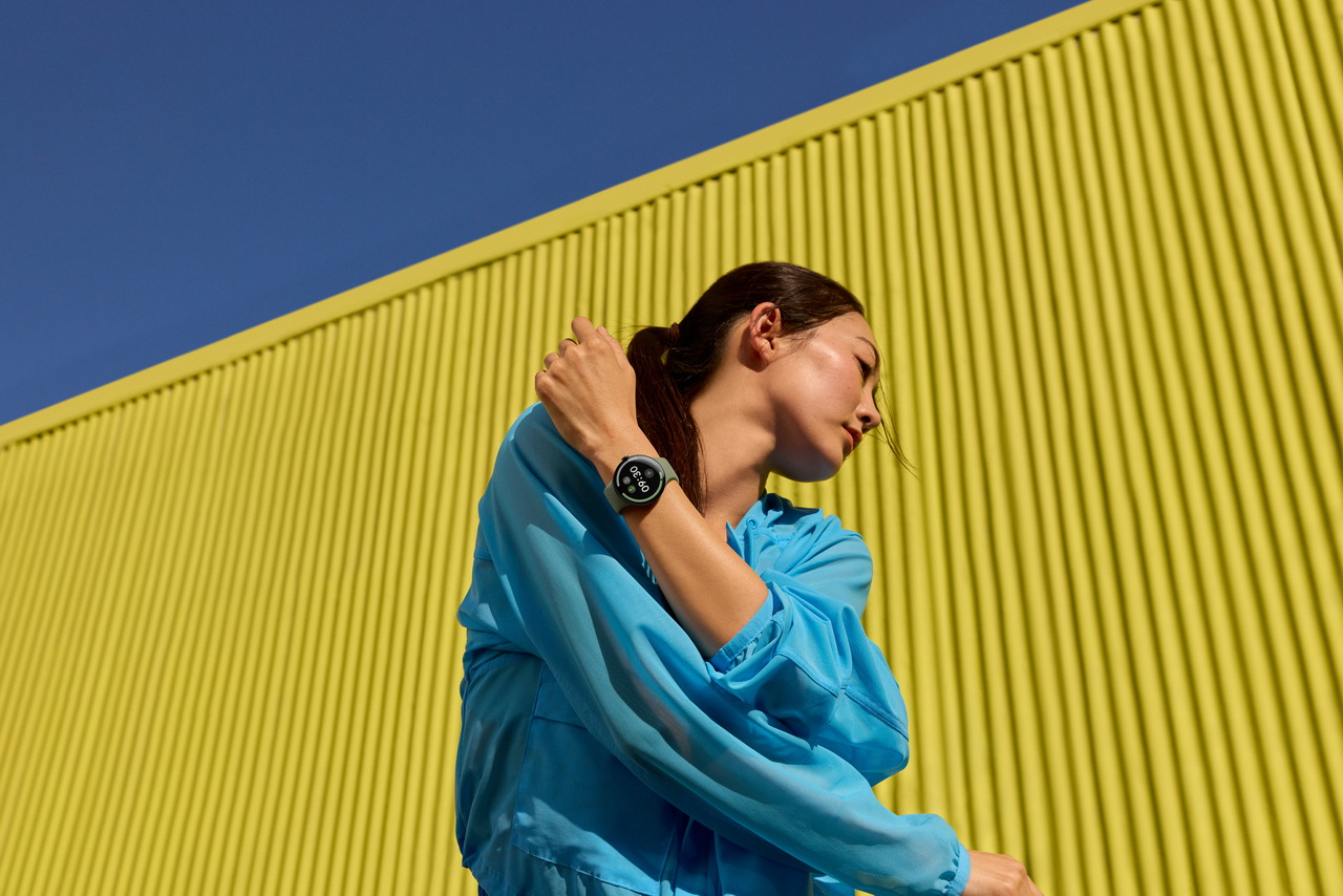 Mujer con sudadera azul y reloj inteligente hace ejercicio frente a pared amarilla en día soleado.
