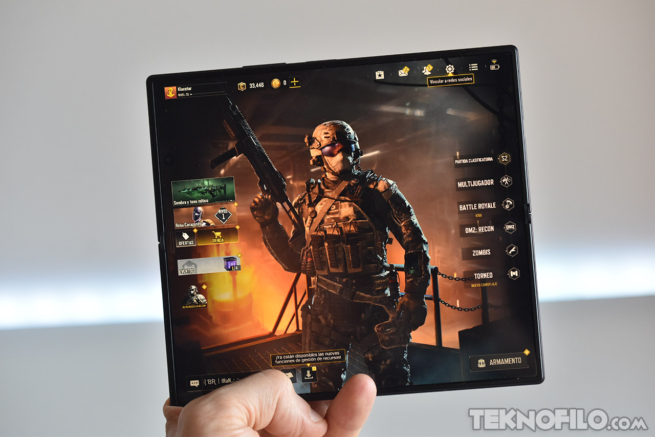 Pantalla de smartphone plegable mostrando un videojuego de disparos, personaje armado con menú de opciones del juego.
