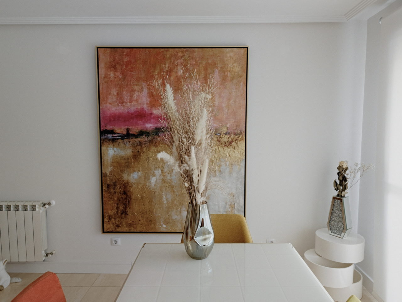 Sala moderna con pintura colorida y jarrón de pampas sobre mesa blanca, decoración interior elegante y minimalista.