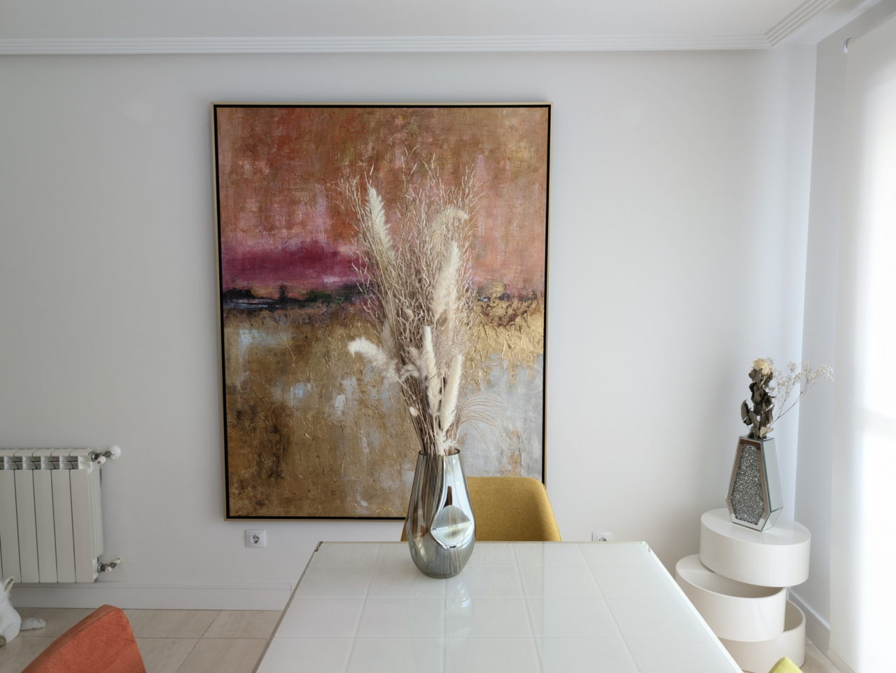 Comedor moderno con pintura abstracta grande, jarrón con ramas decorativas y muebles blancos minimalistas.