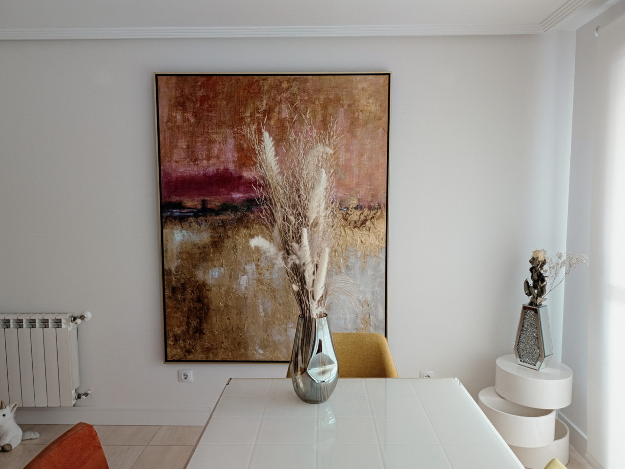 Cuadro abstracto en sala moderna con decoración de jarrones y pampas sobre mesa blanca.