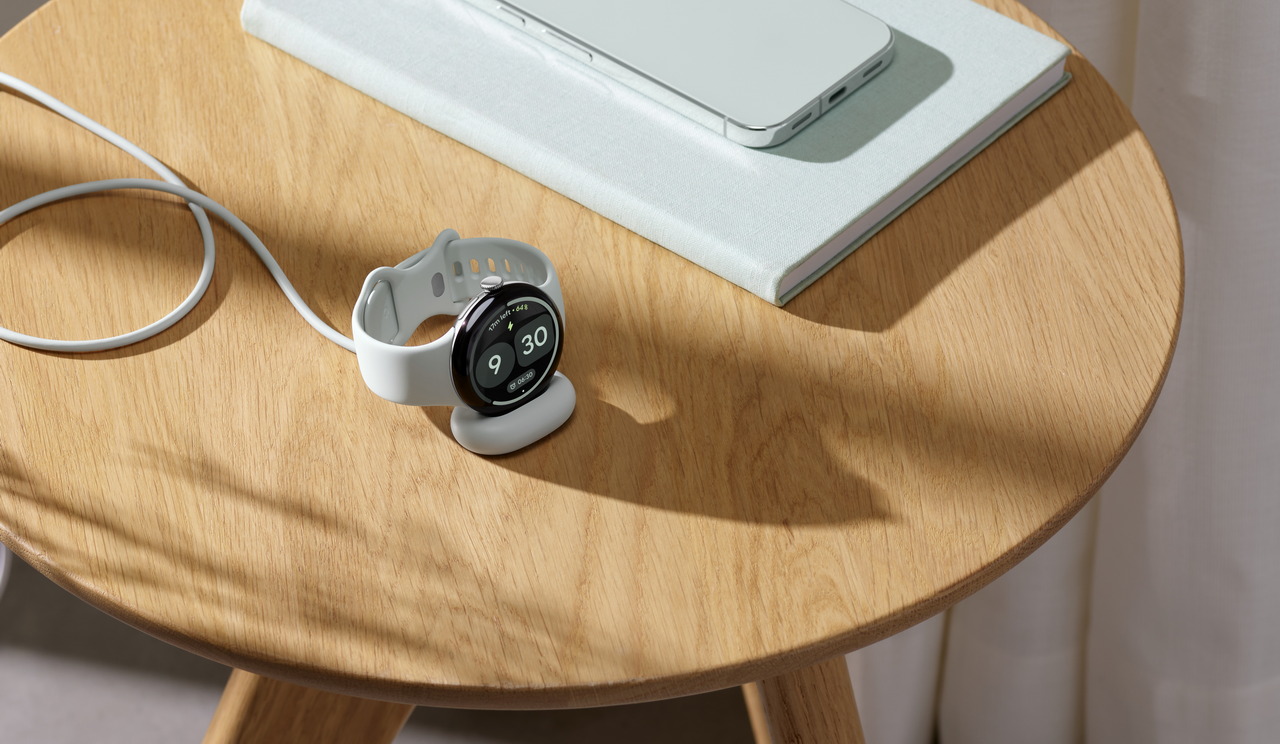 Reloj inteligente cargándose en una mesa de madera junto a un cuaderno y un smartphone gris.