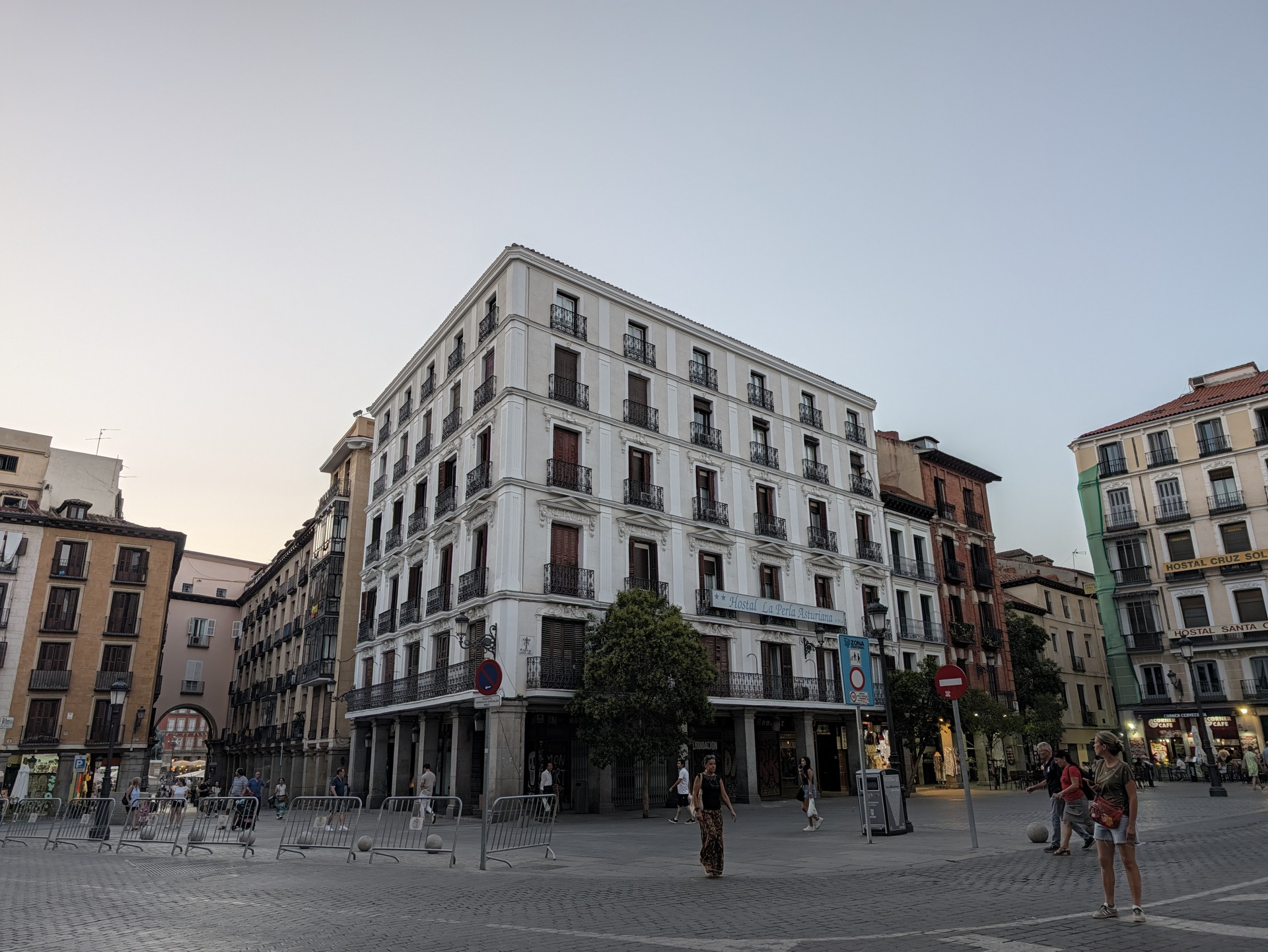 Edificios históricos en una plaza de Madrid al atardecer, con personas caminando por la calle y cielo despejado.