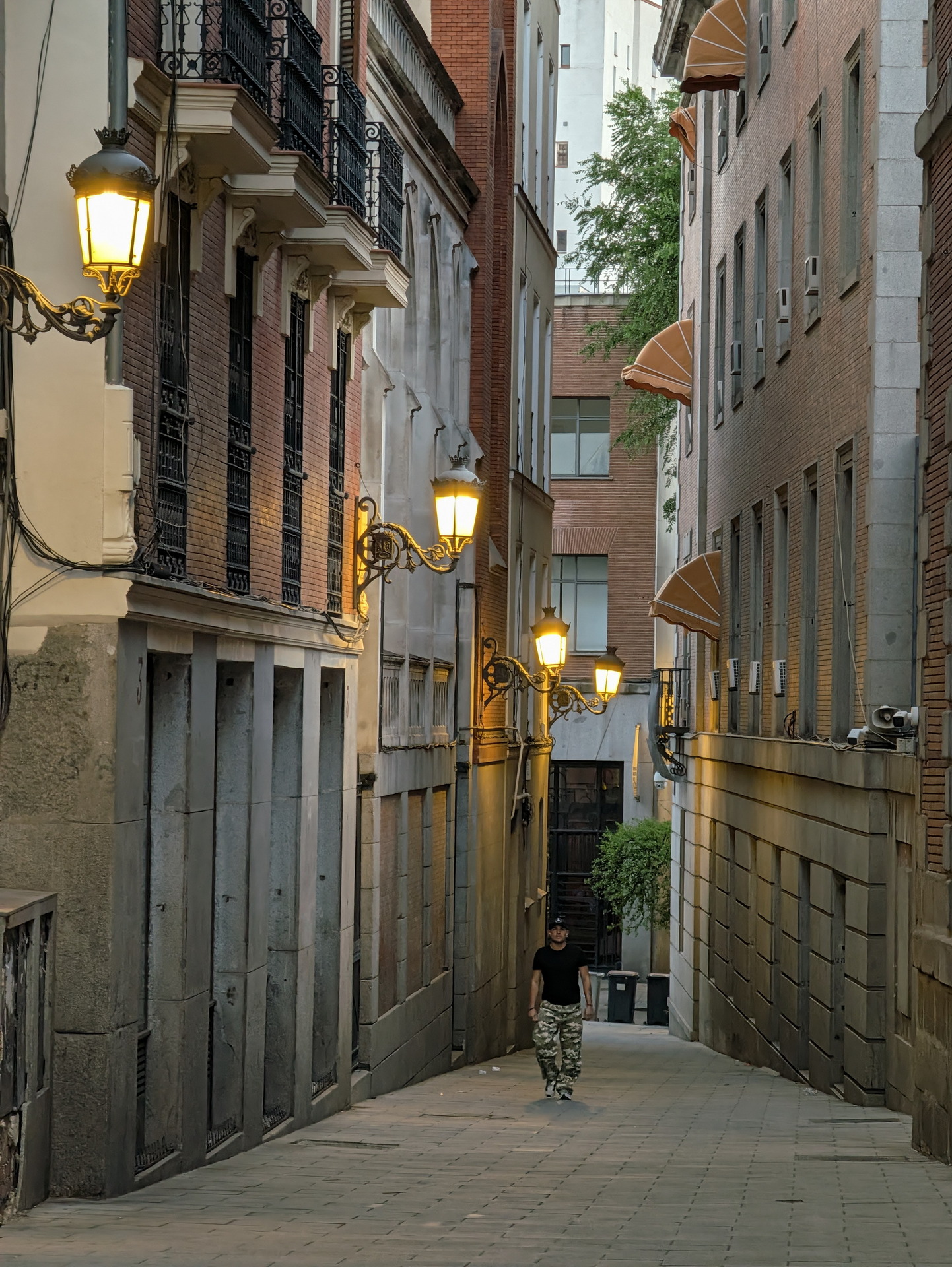 Callejón estrecho en ciudad europea con farolas encendidas y hombre caminando al atardecer.