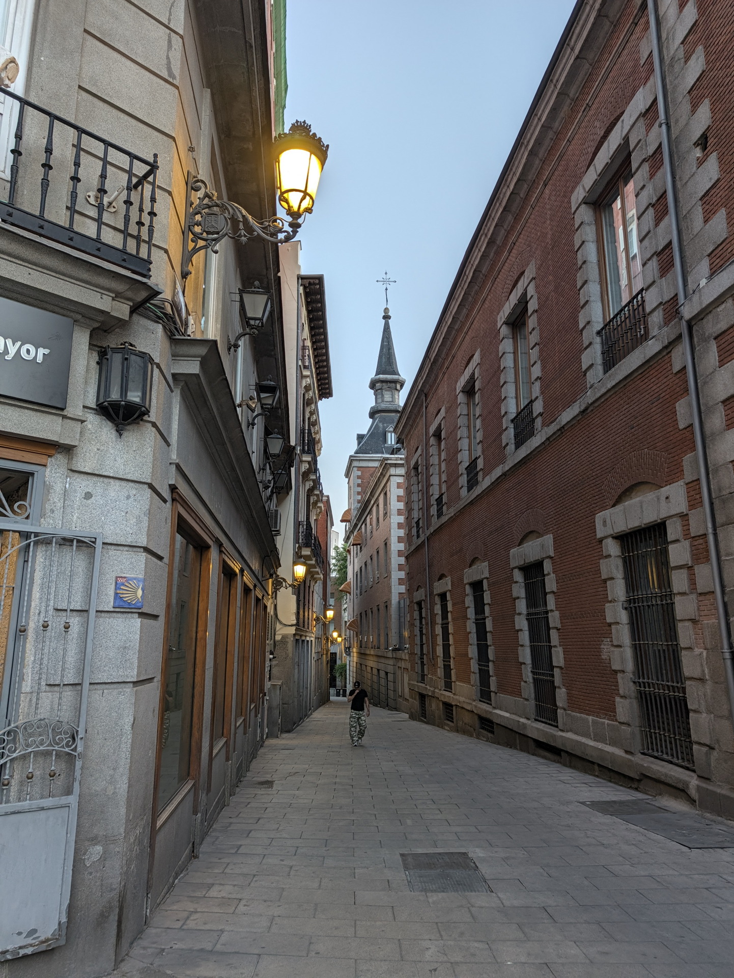 Calle estrecha en Madrid al atardecer con farolas encendidas y edificio histórico al fondo.