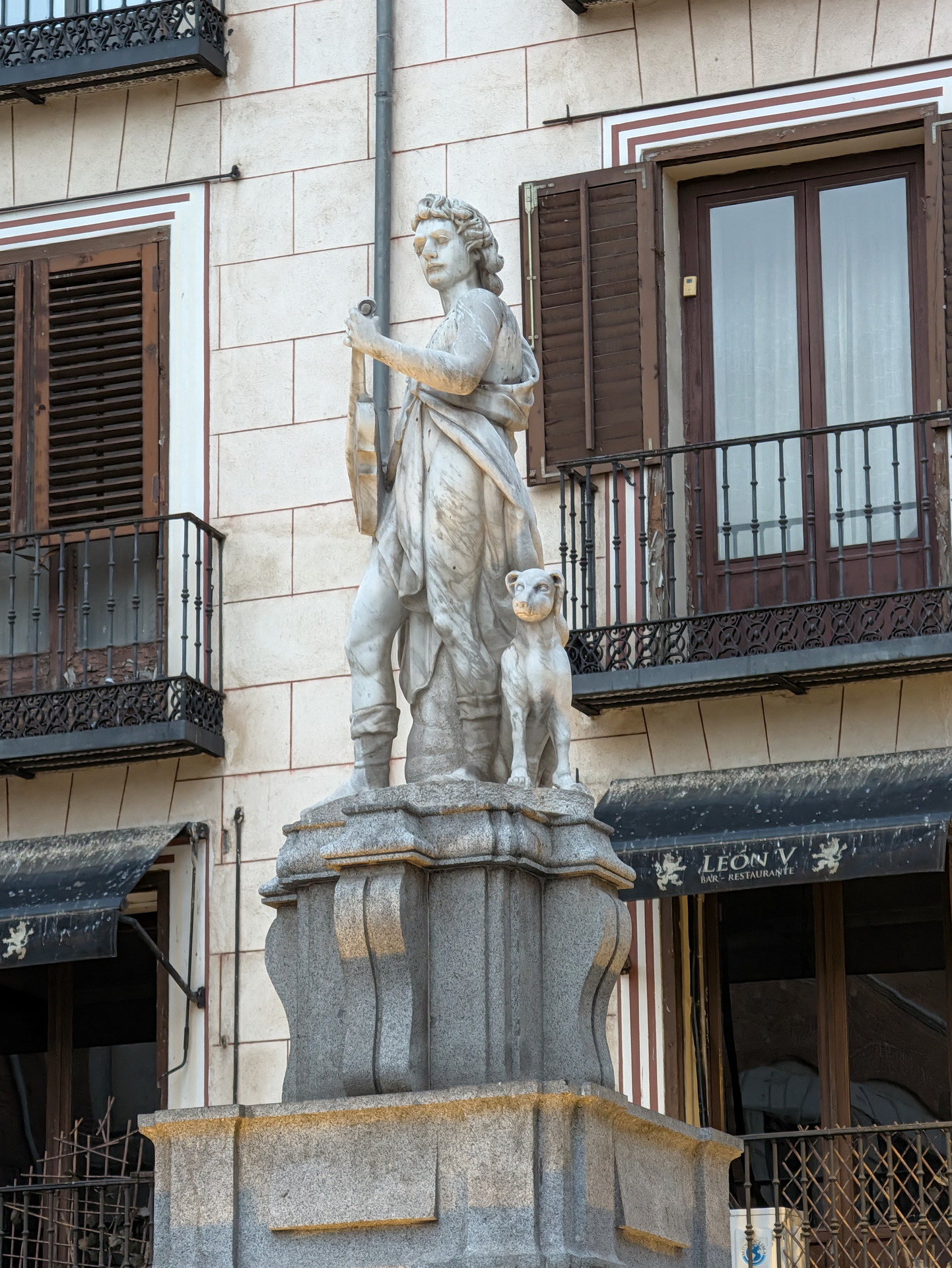 Estatua clásica en una plaza, representando una figura con instrumento musical y un perro al lado, en un entorno urbano.