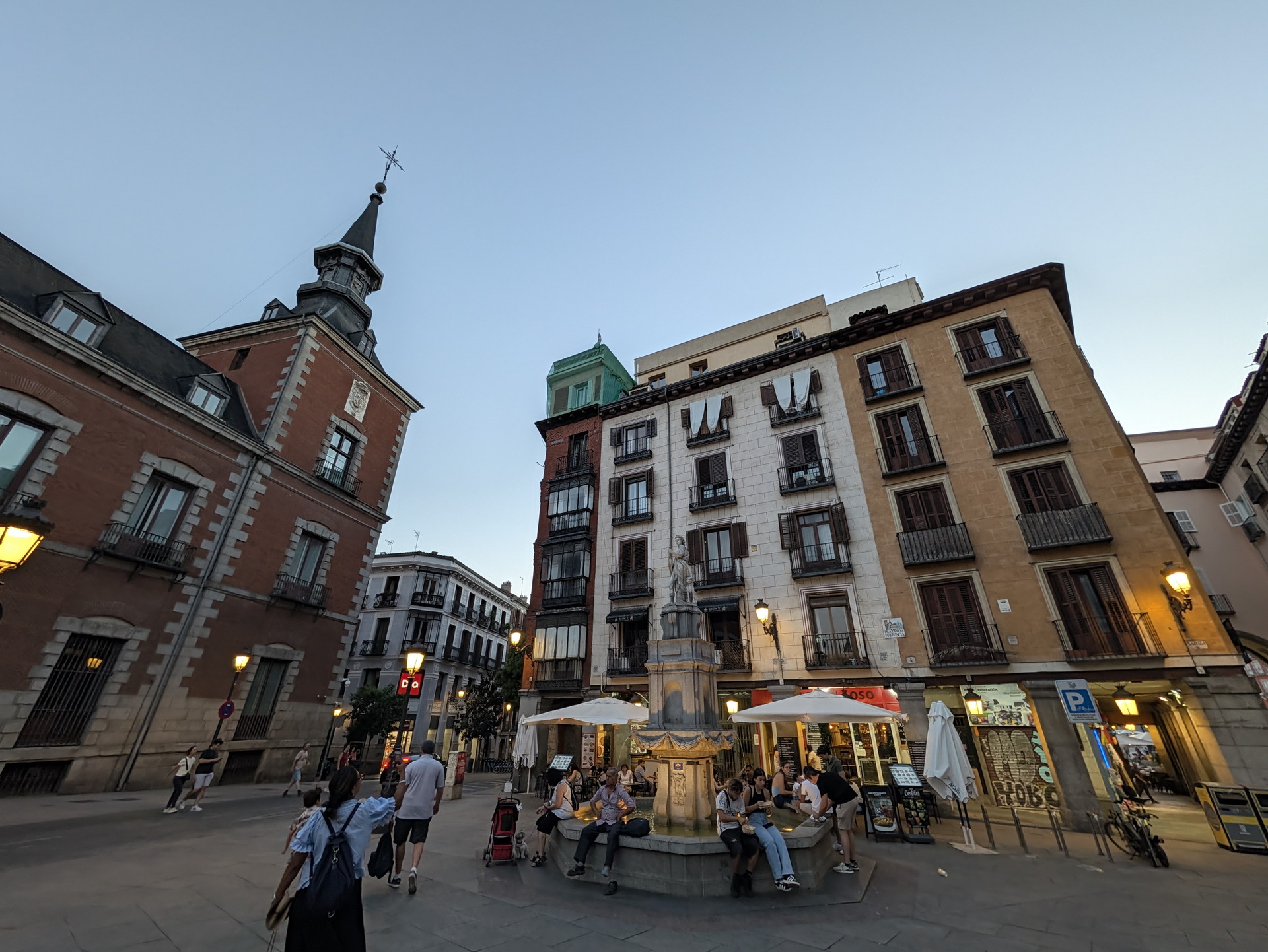 Plaza en Madrid al anochecer, con edificios históricos y personas disfrutando cerca de la fuente central.