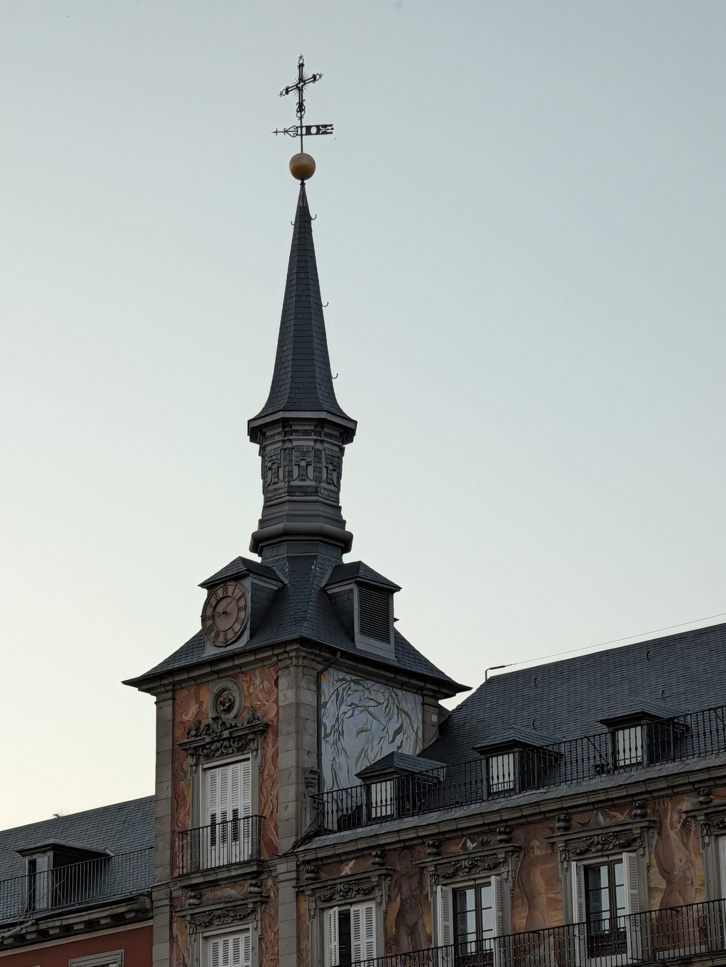 Torre de reloj con tejado puntiagudo en edificio histórico al atardecer, decorado con murales y ventanas con balcones.