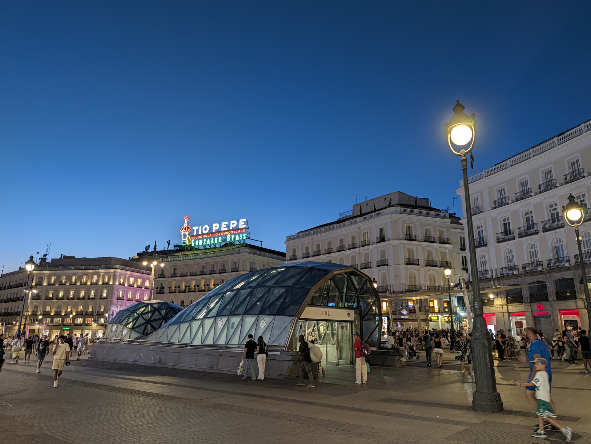Entrada al metro en Puerta del Sol, Madrid, al anochecer, con gente paseando y letrero de Tío Pepe iluminado.
