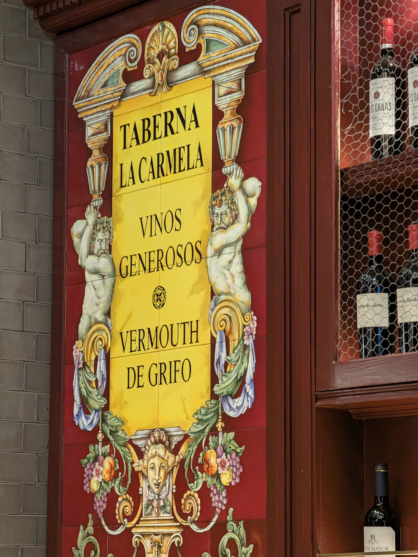 Cartel colorido de Taberna La Carmela destacando vinos generosos y vermut de grifo, con botellas de vino al lado.