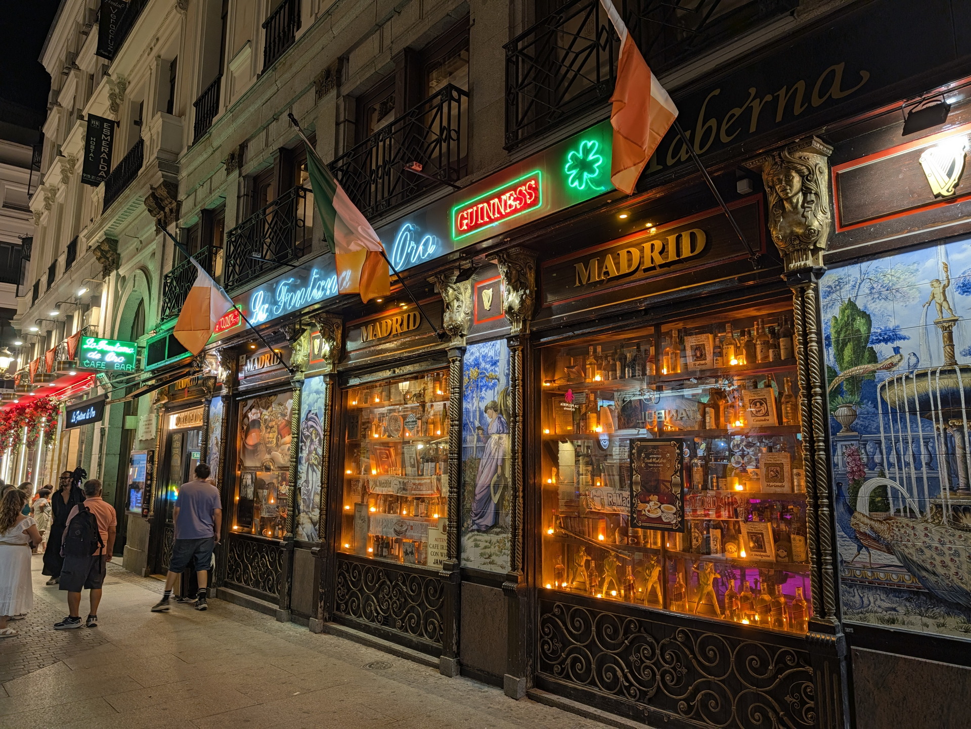 Fachada de taberna irlandesa iluminada en Madrid con banderas y vitrinas decoradas, ambiente nocturno animado.