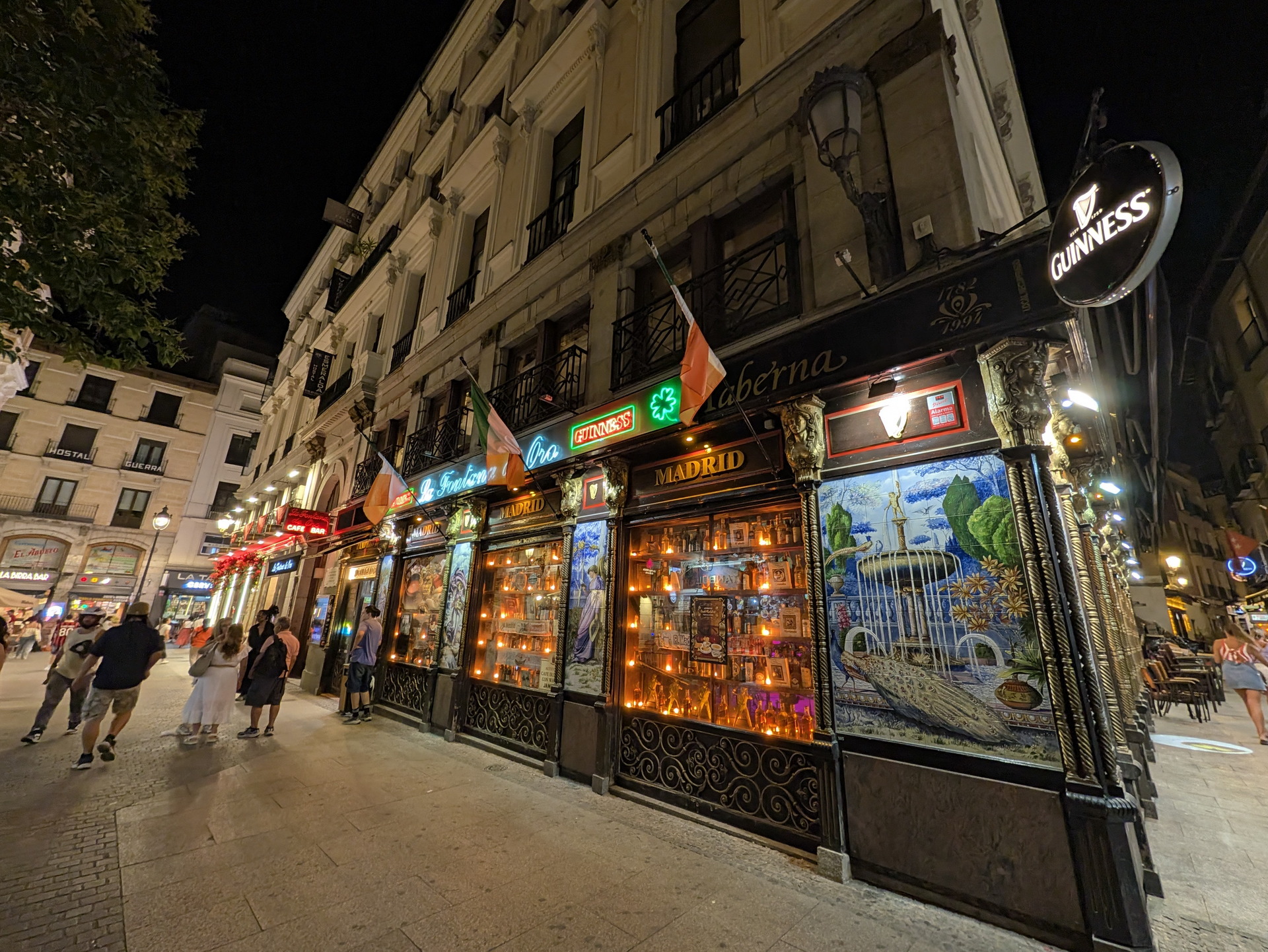 Una taberna iluminada por la noche en una calle concurrida de Madrid, con gente caminando y charlando.