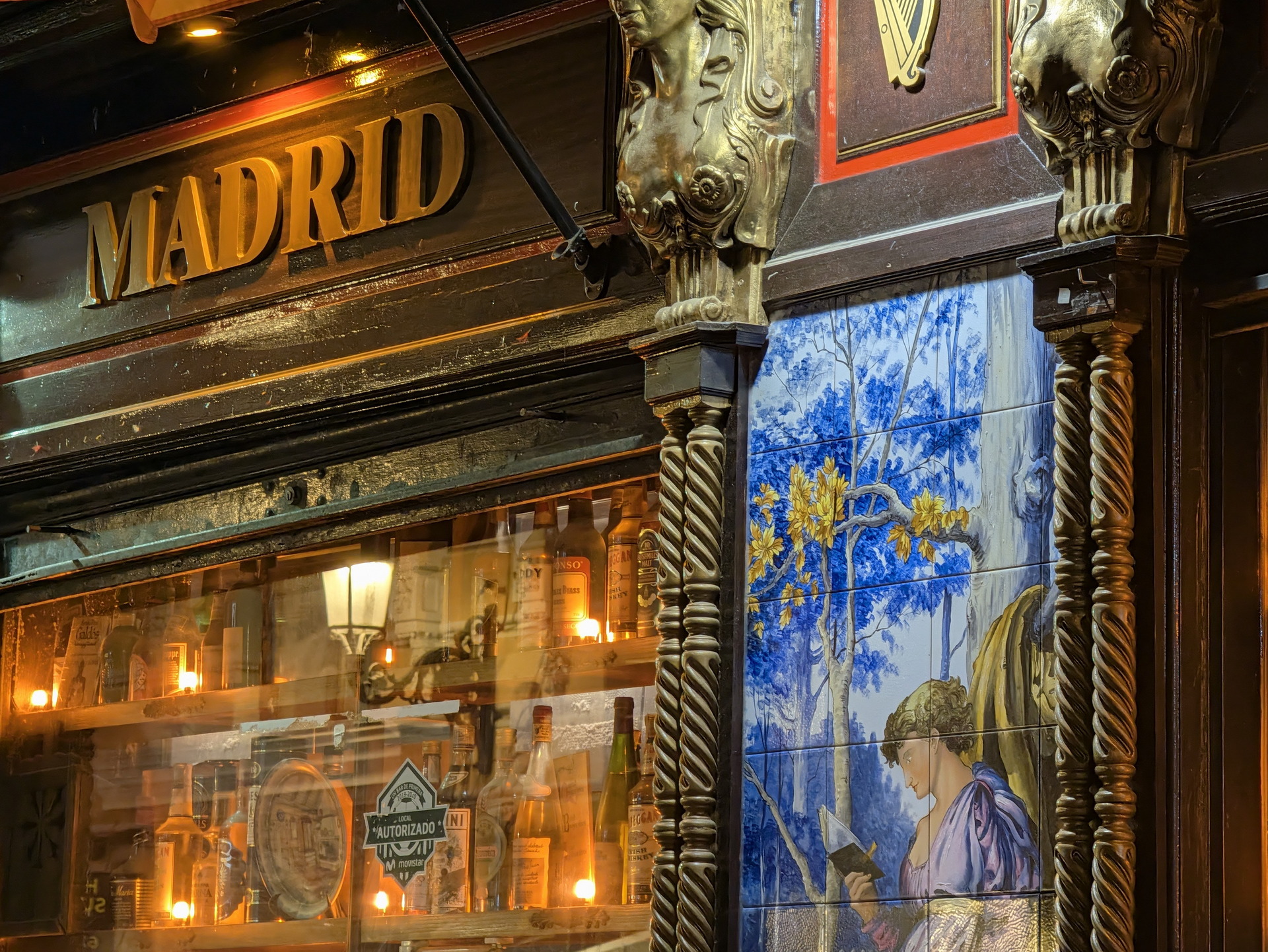 Fachada de un bar en Madrid con vidrieras iluminadas y mosaico artístico de una figura femenina entre árboles.