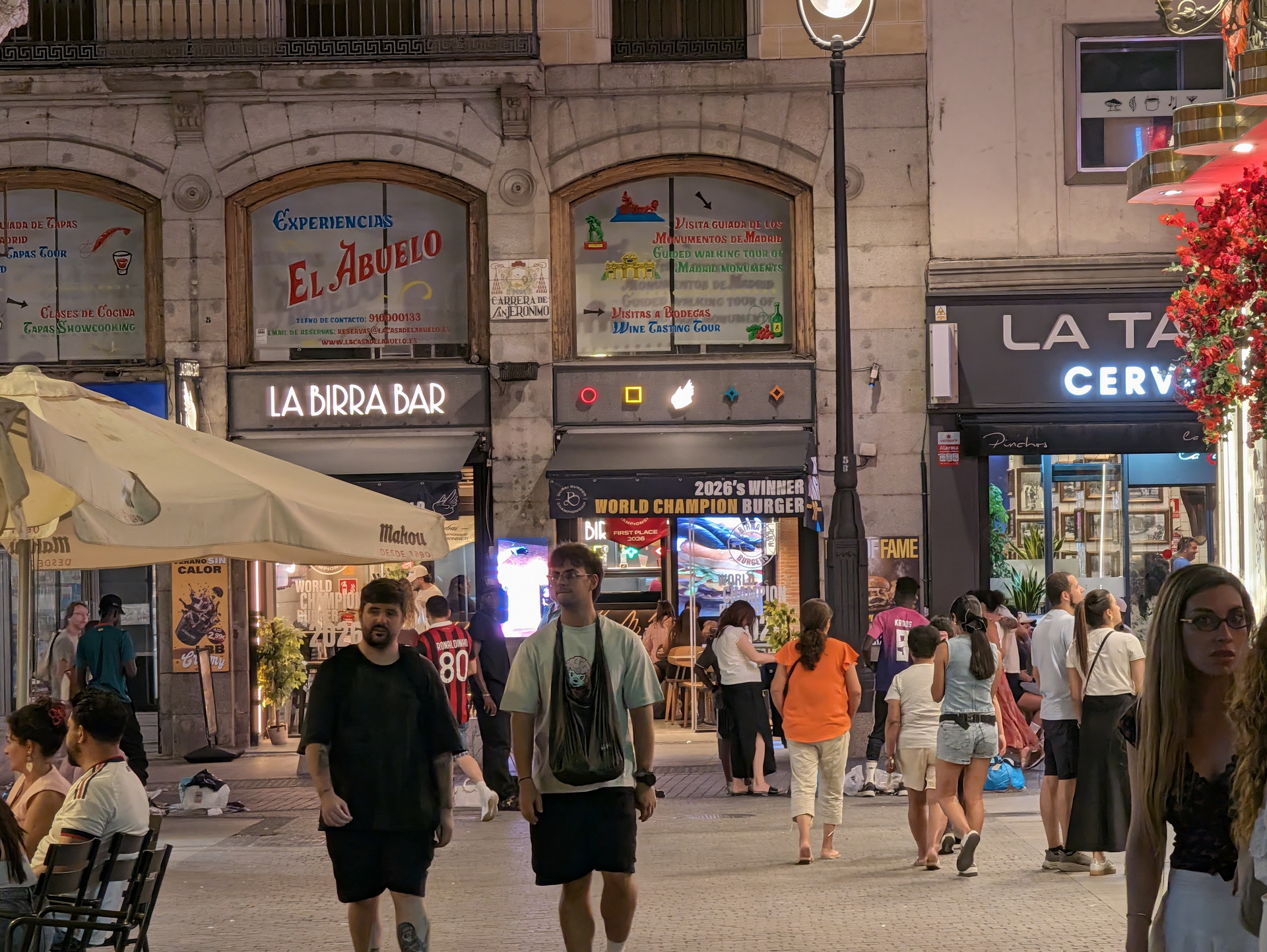 Calle transitada en la noche con bares y restaurantes iluminados, gente disfrutando en terrazas al aire libre.