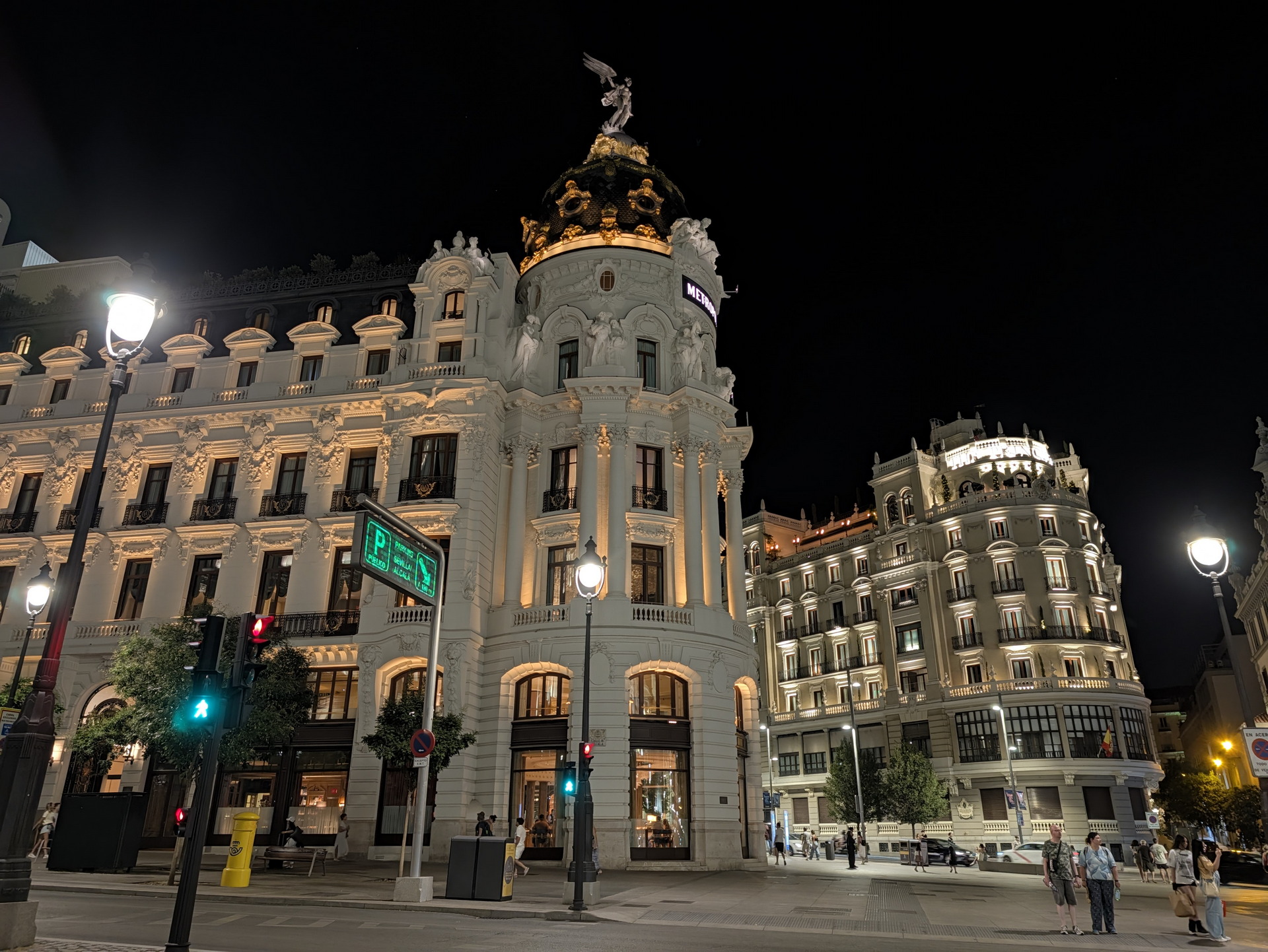 Edificio histórico iluminado en una avenida de Madrid, con cielo nocturno y transeúntes paseando.