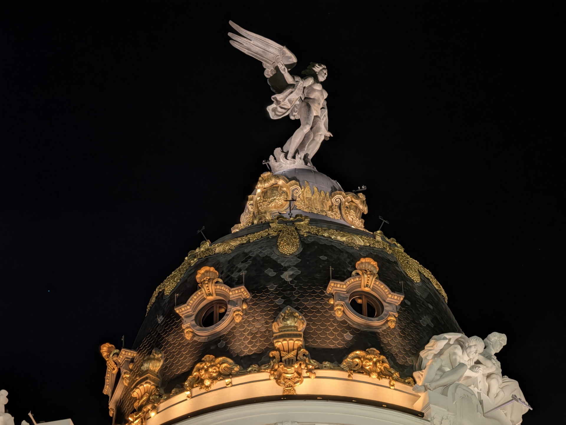 Cúpula del Edificio Metropolis en Madrid iluminada de noche con una escultura alada en la cima.