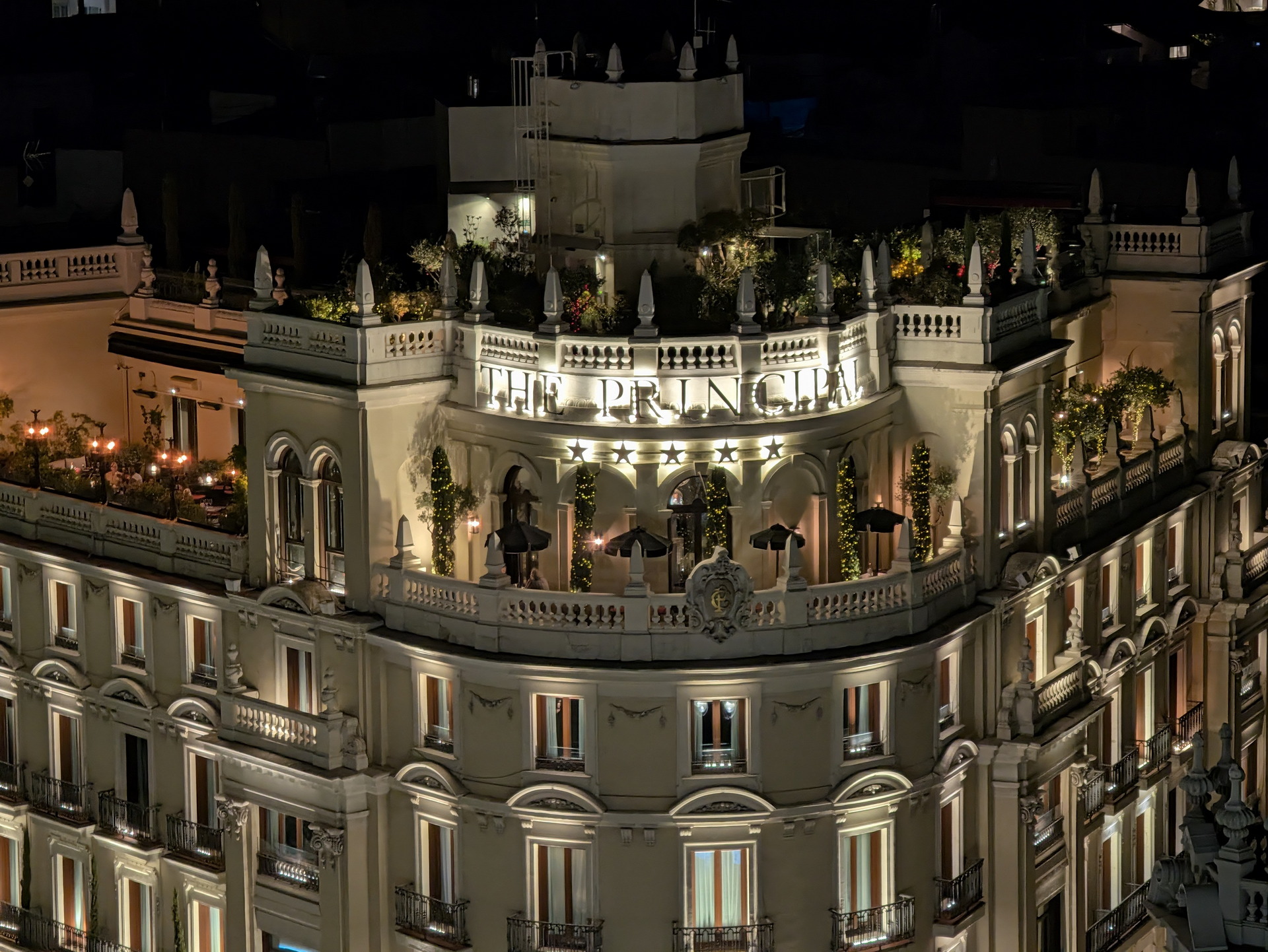 Edificio iluminado por la noche con terraza y decoración elegante en la fachada superior, rodeado de vegetación.