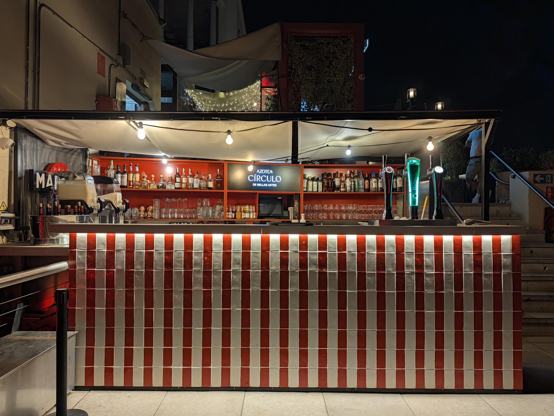 Bar nocturno con luces cálidas en azotea, estantería de licores y decorado con azulejos rojos y blancos.
