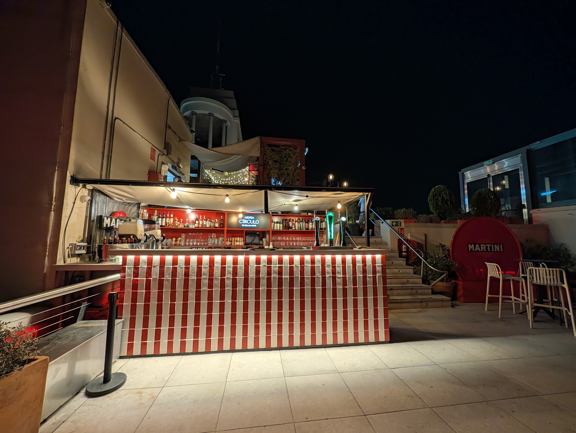 Bar al aire libre con luces y decoración roja y blanca en una terraza nocturna. Ambiente acogedor y moderno.