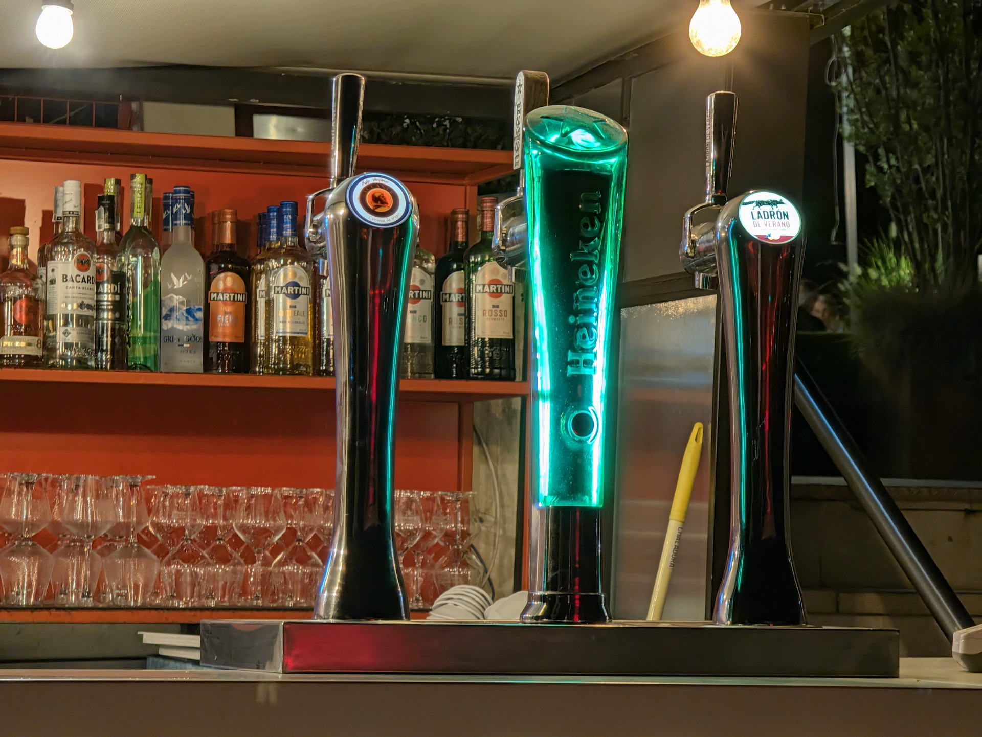 Grifos de cerveza en un bar con botellas de licor y vasos alineados en el fondo, ambiente nocturno y relajado.