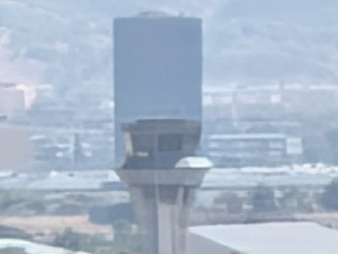 Torre de control de aeropuerto con edificios y montañas en el fondo, vista desde lejos.