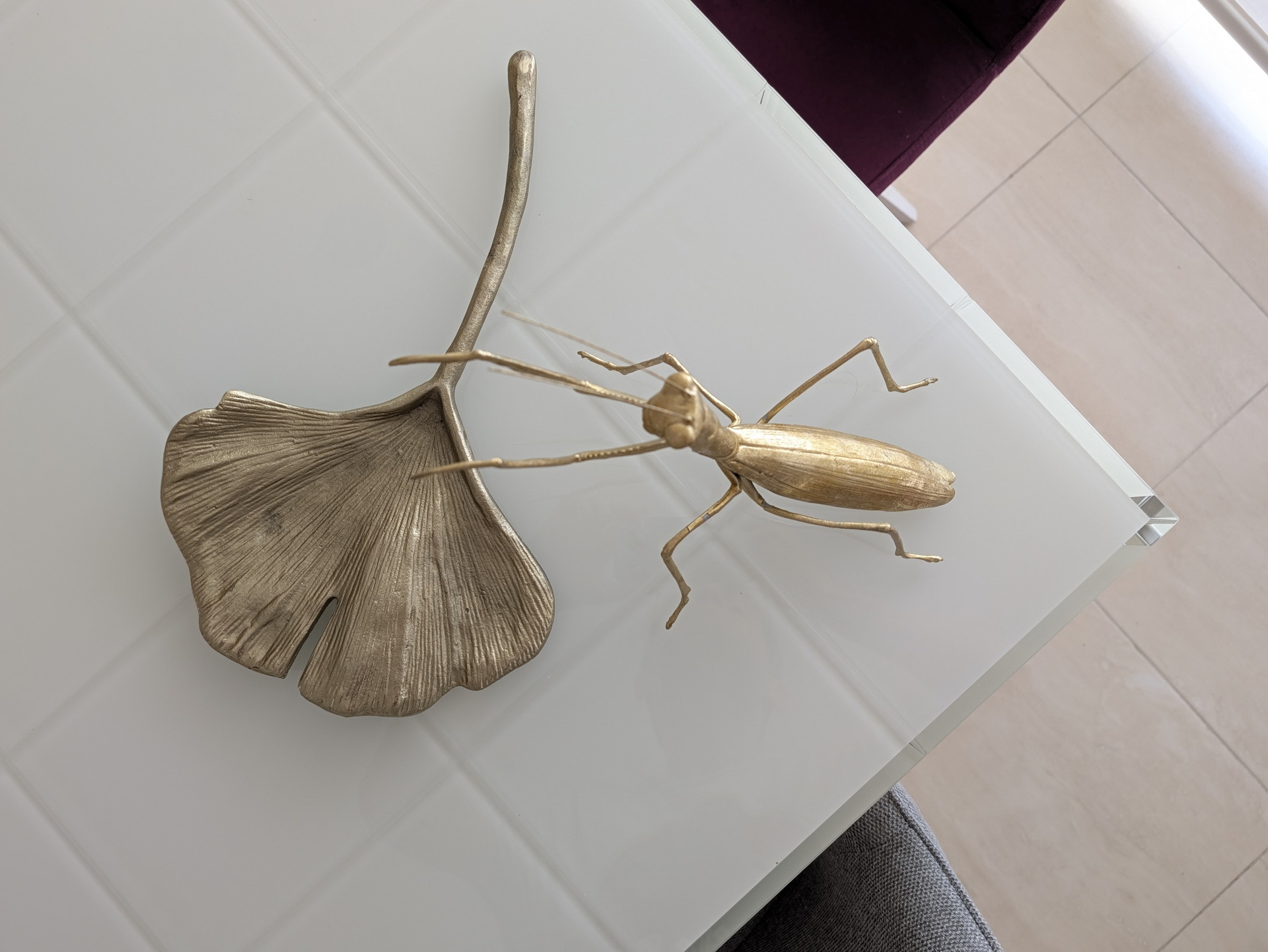 Escultura dorada de hoja de Ginkgo y mantis sobre mesa blanca minimalista en ambiente moderno.
