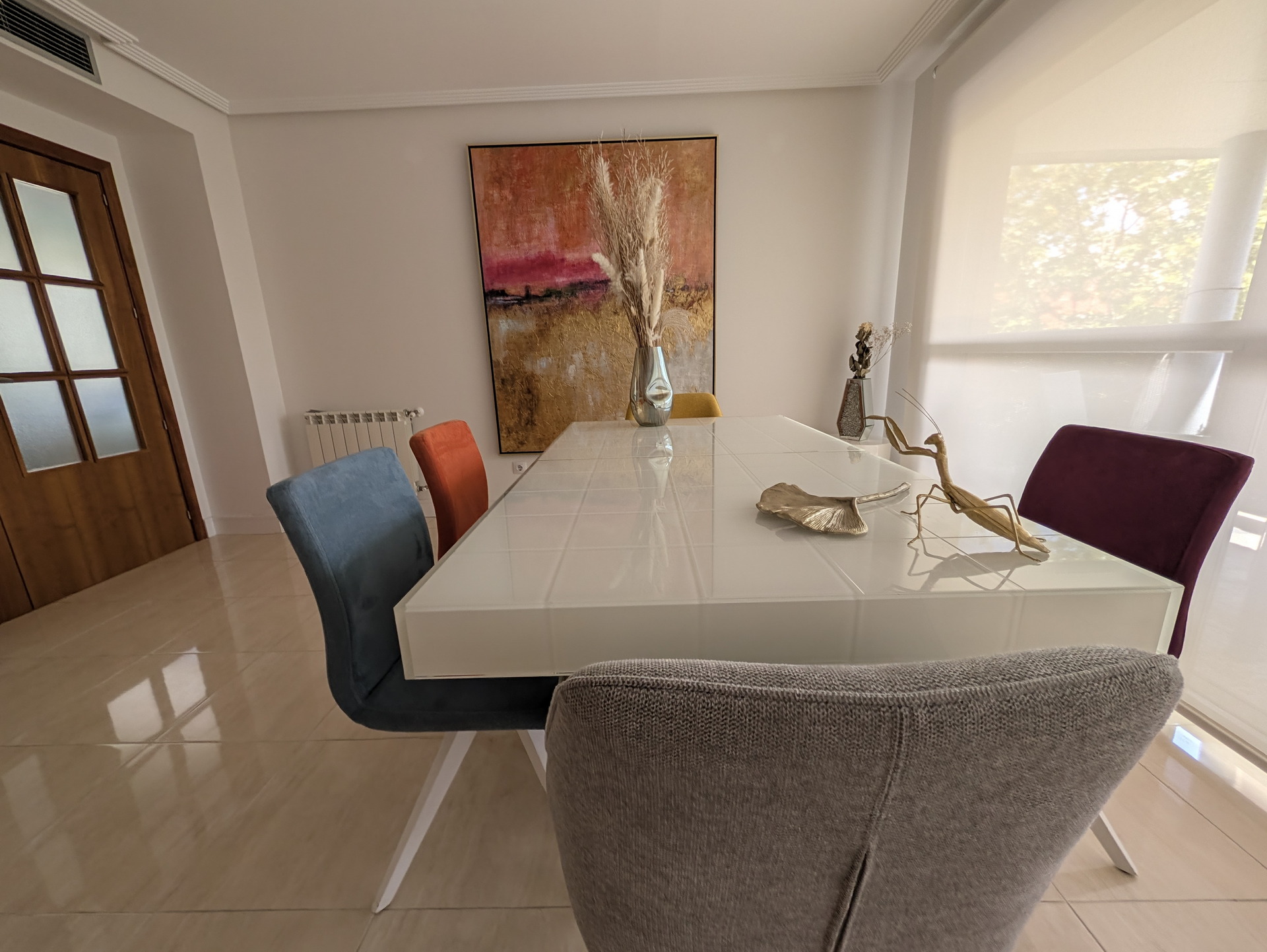 Comedor moderno con mesa blanca, sillas de colores y decoración minimalista junto a gran ventana.