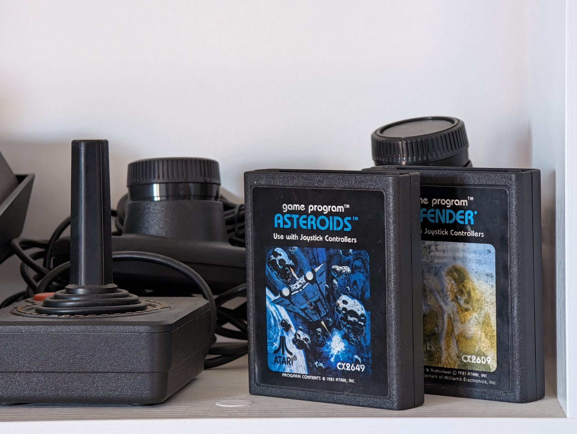 Cartuchos retro de Atari Asteroids y Defender con controladores de joystick en estante blanco.