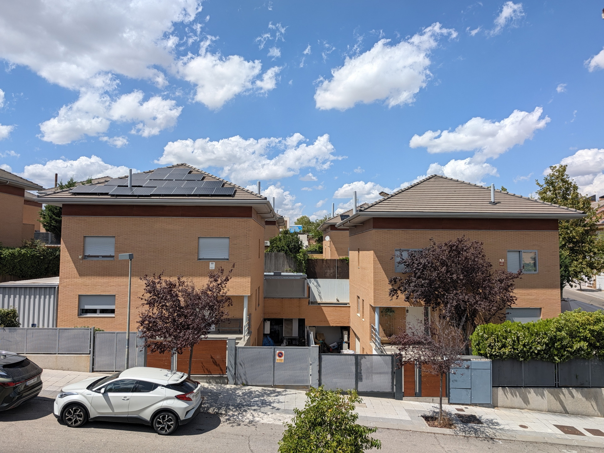 Dos casas modernas con paneles solares en tejados, cielo azul y coches aparcados enfrente.