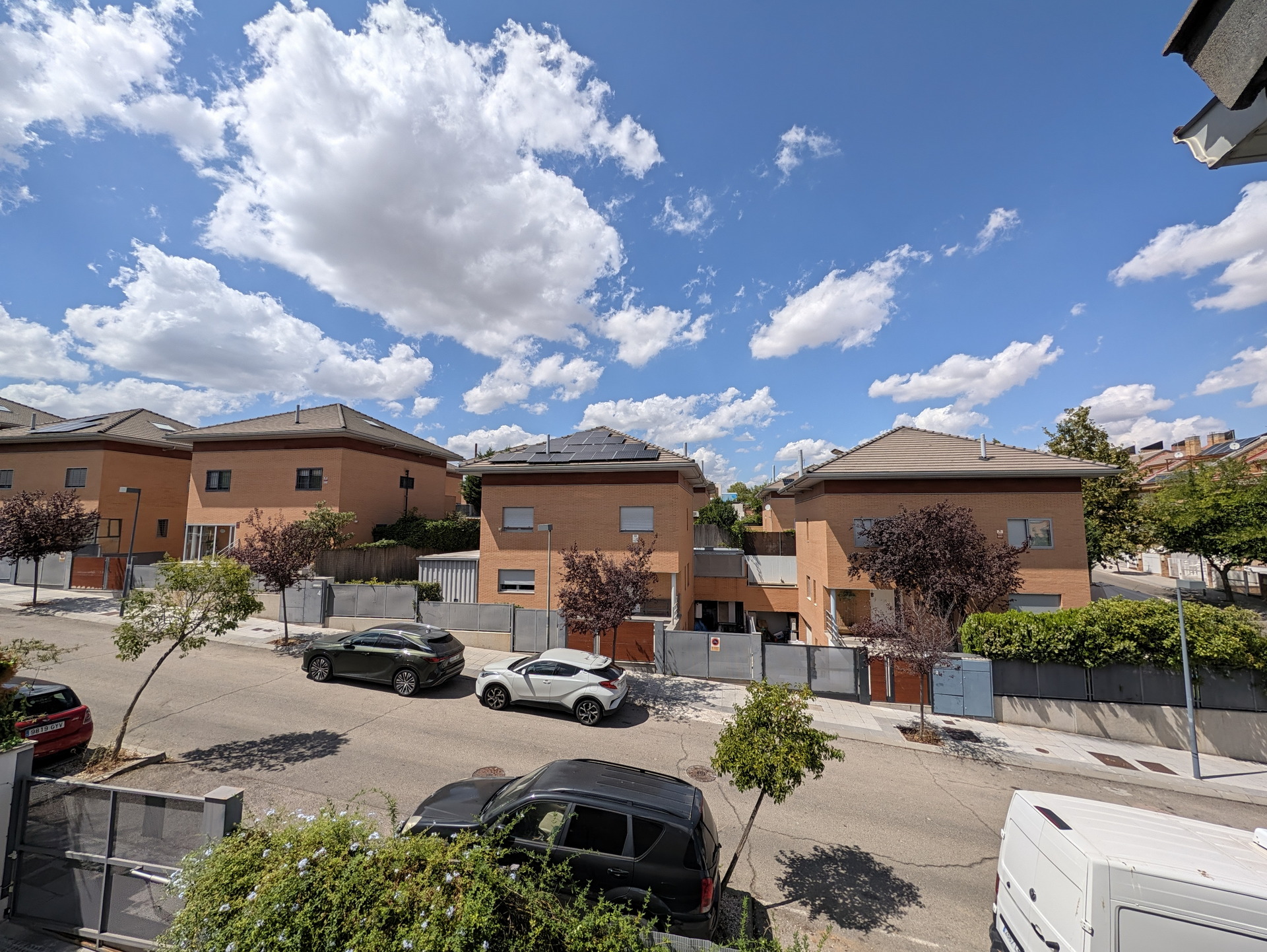 Calle residencial con casas de ladrillo y árboles en día soleado, cielo azul con nubes esponjosas.