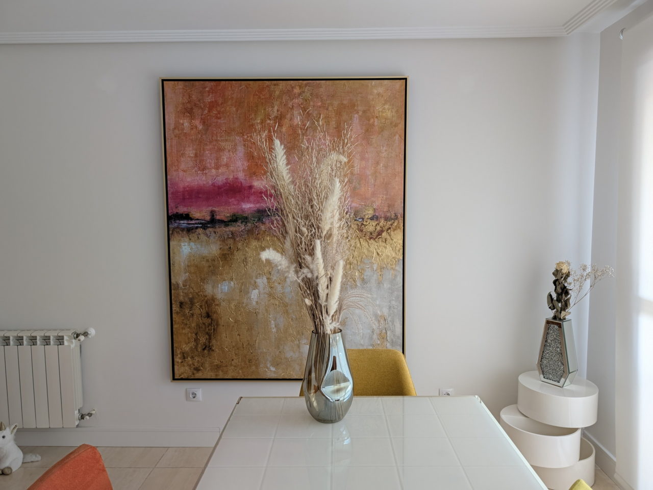 Pintura abstracta en pared blanca junto a jarrón con flores secas en mesa de comedor moderno. Decoración minimalista.