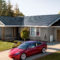 Casa con techo solar y auto eléctrico rojo en la entrada, rodeados de árboles y jardín verde. Innovación sostenible.