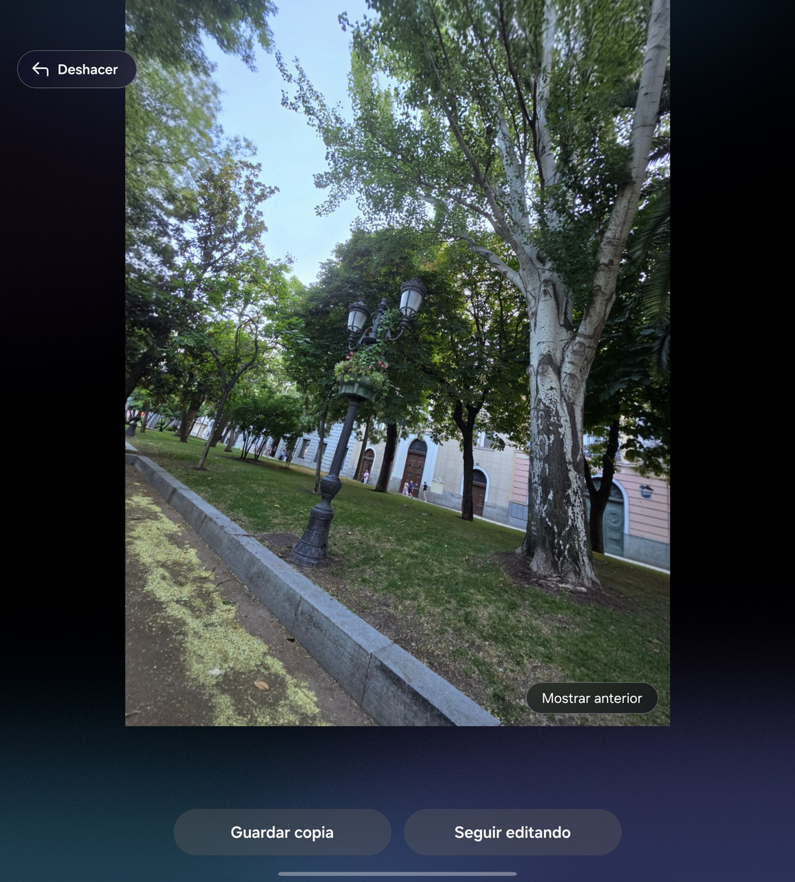 Paseo arbolado con farol antiguo, césped verde y edificios históricos al fondo bajo un cielo azul claro.
