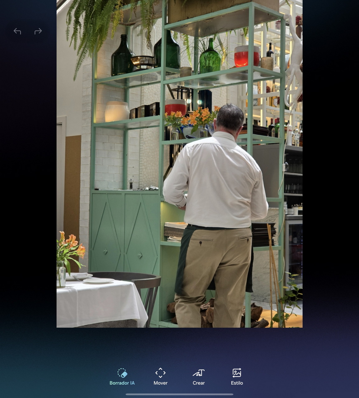 Camarero en restaurante organizando estantería decorativa con botellas y plantas colgantes. Ambiente acogedor.