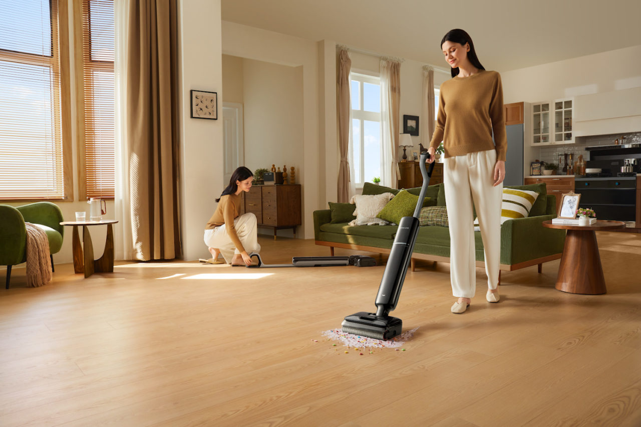 Mujer aspirando suelo de sala moderna, luz natural, sofá verde y decoración elegante, limpieza hogar eficiente.