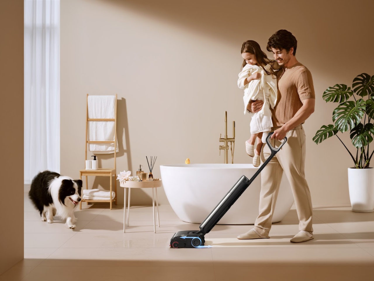 Padre limpiando el suelo con aspiradora, sostiene a su hija, mientras un perro observa en un baño moderno y luminoso.