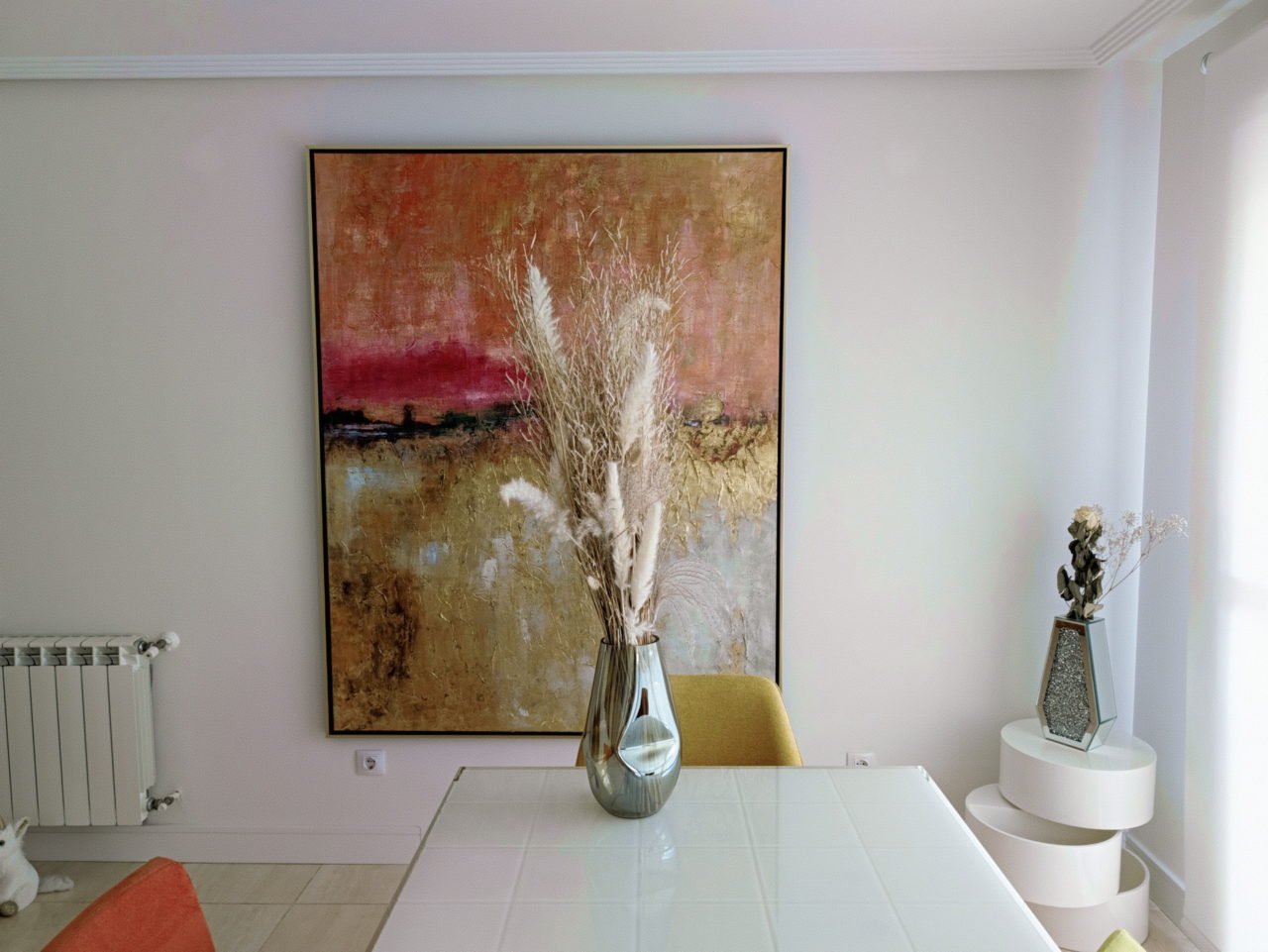 Pintura abstracta colorida en pared de salón moderno, con jarrón de flores secas sobre mesa blanca.
