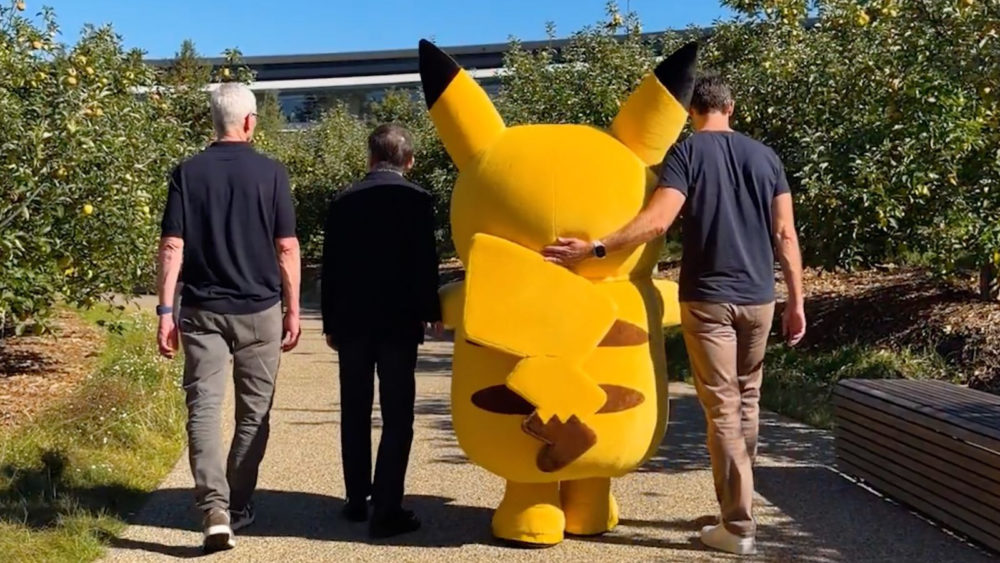 Un Pikachu gigante caminando con tres personas en un jardín soleado.