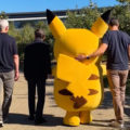 Un Pikachu gigante caminando con tres personas en un jardín soleado.