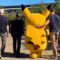 Un Pikachu gigante caminando con tres personas en un jardín soleado.