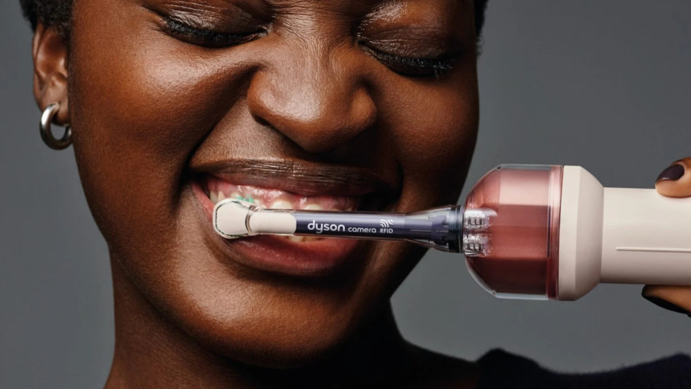 Dyson lanza un cepillo de dientes con cámara e IA que cuesta 479 euros Mujer cepillándose los dientes con un cepillo eléctrico Dyson, mostrando una sonrisa saludable.
