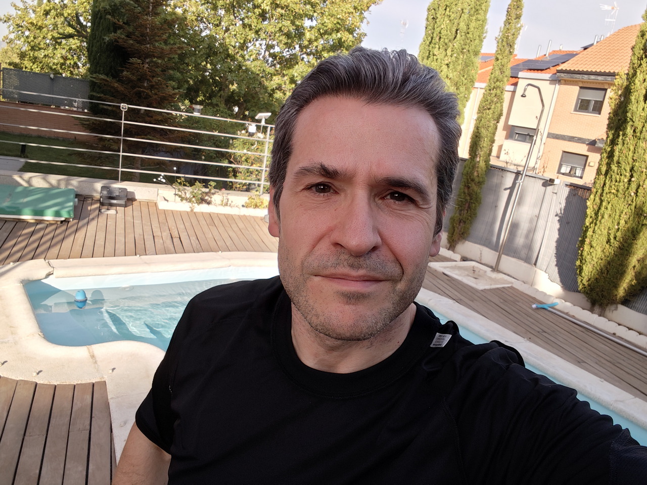 Hombre con camiseta negra junto a una piscina en un patio, rodeado de árboles y una valla.